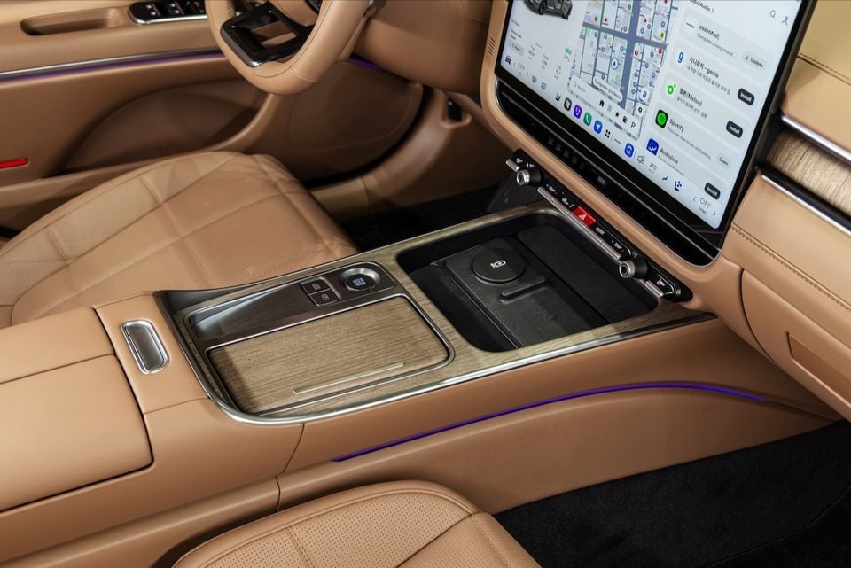 Interior view of the 2027 Hyundai Grandeur Pleos showing the centre console with wood trim, wireless charging pad, and a large touchscreen infotainment display with navigation and apps visible, surrounded by tan leather seats and purple ambient lighting.