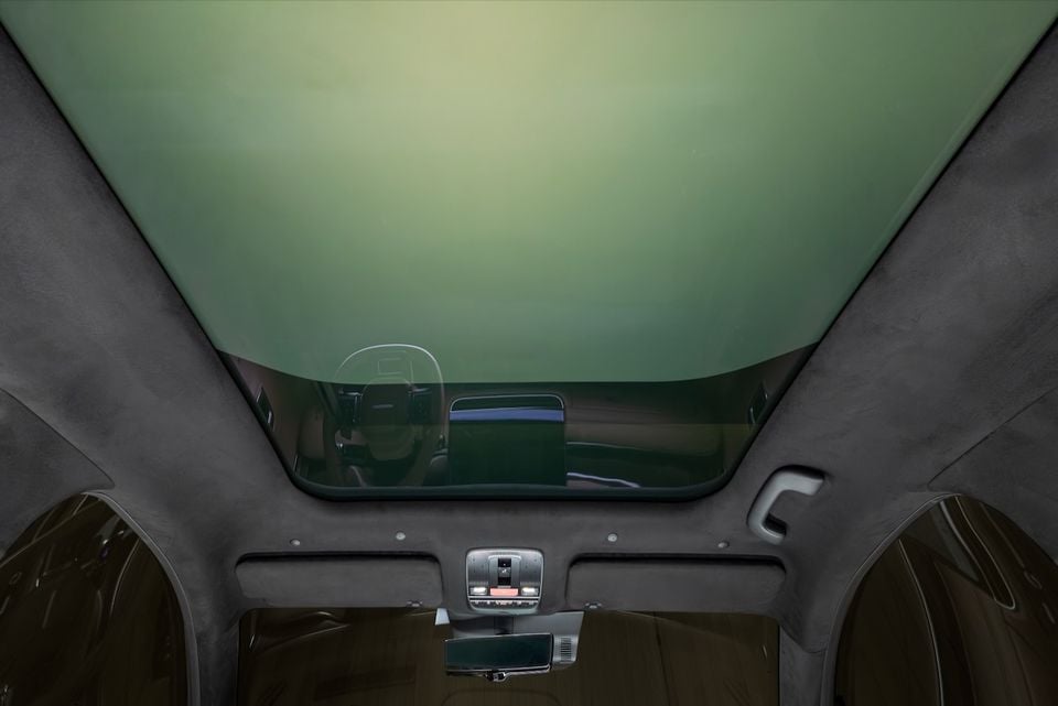 Interior view of a car looking up at a large panoramic glass roof with green tinting, showing the headliner, overhead console with reading lights, and rearview mirror, with the dashboard and steering wheel visible through the glass partition.