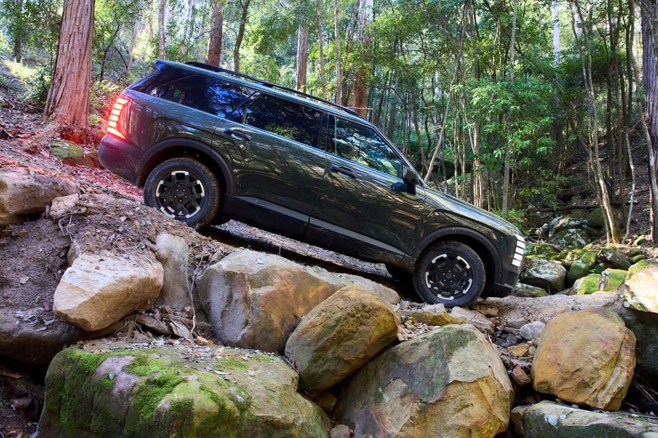 A dark green 2027 Hyundai Palisade XRT Pro SUV navigating a rocky off-road trail in a forest, seen from the side.