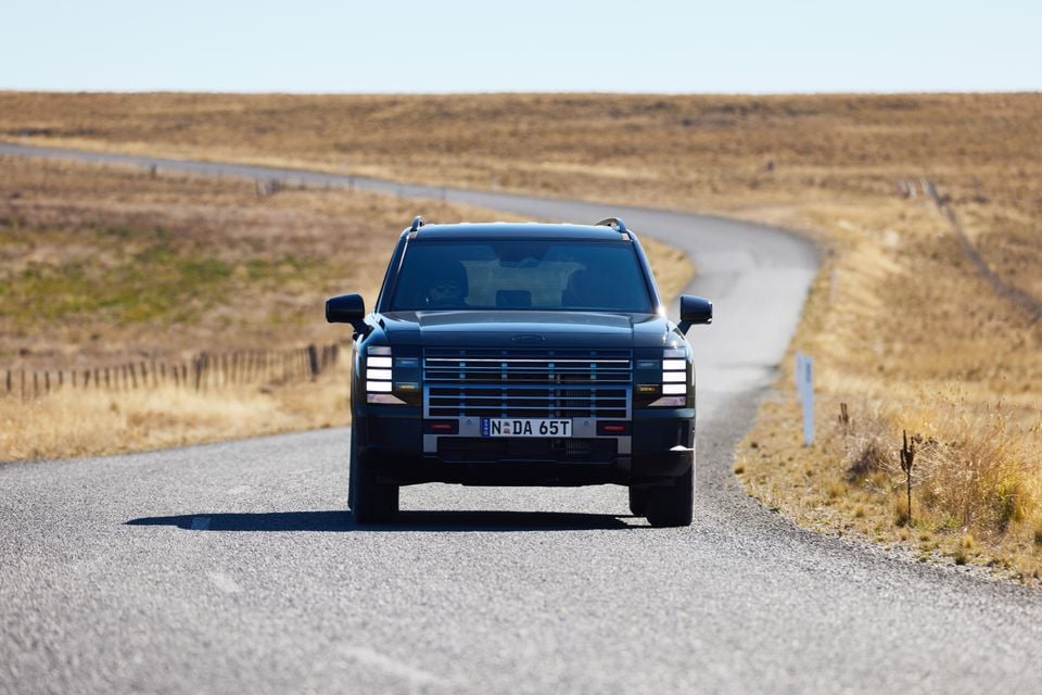 A dark blue 2027 Hyundai Palisade XRT Pro SUV seen from the front, driving along a winding rural road surrounded by dry grassland in Australia.
