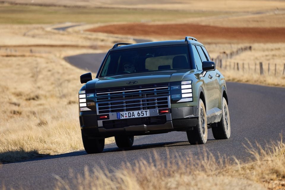 A dark green Hyundai Palisade XRT Pro SUV driving along a rural road, seen from the front at a slight angle, with dry grassland in the background