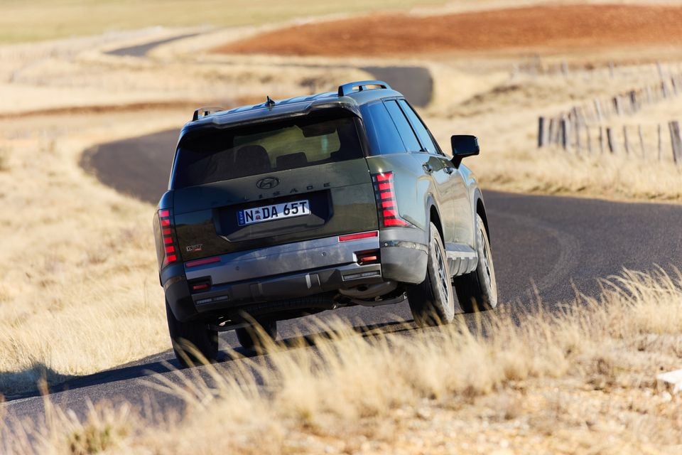 Rear view of a Hyundai Palisade XRT Pro SUV driving along a winding rural road surrounded by dry grassland