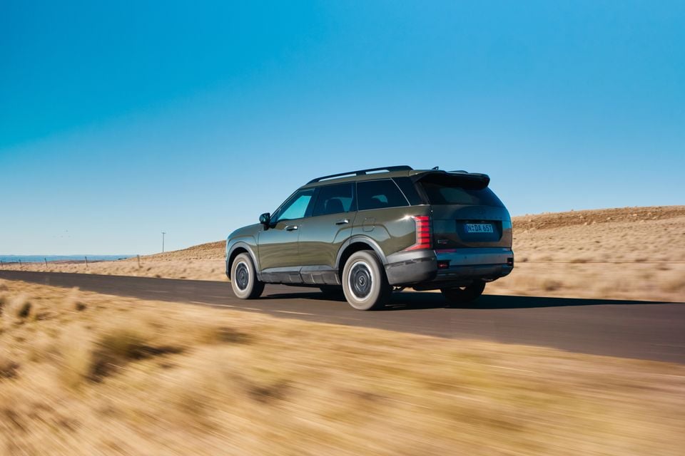 A dark green 2027 Hyundai Palisade XRT Pro SUV driving on a rural road, seen from the rear three-quarter angle against a clear blue sky.