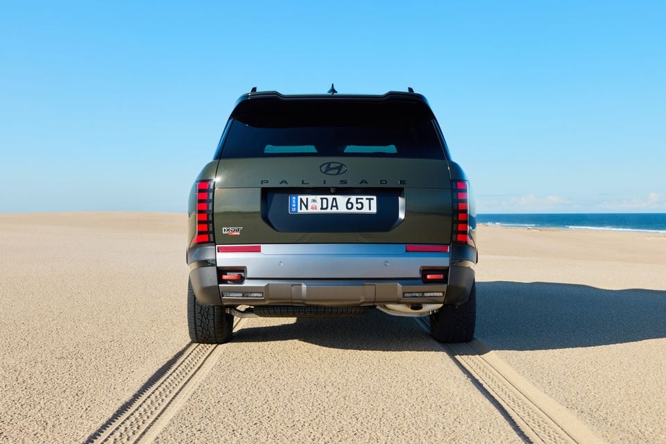Rear view of a dark green 2027 Hyundai Palisade X-RT Pro SUV parked on a sandy beach with the ocean visible in the background