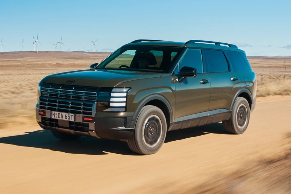 A dark green 2027 Hyundai Palisade XRT Pro large SUV driving on a dirt road in an arid landscape with wind turbines in the background