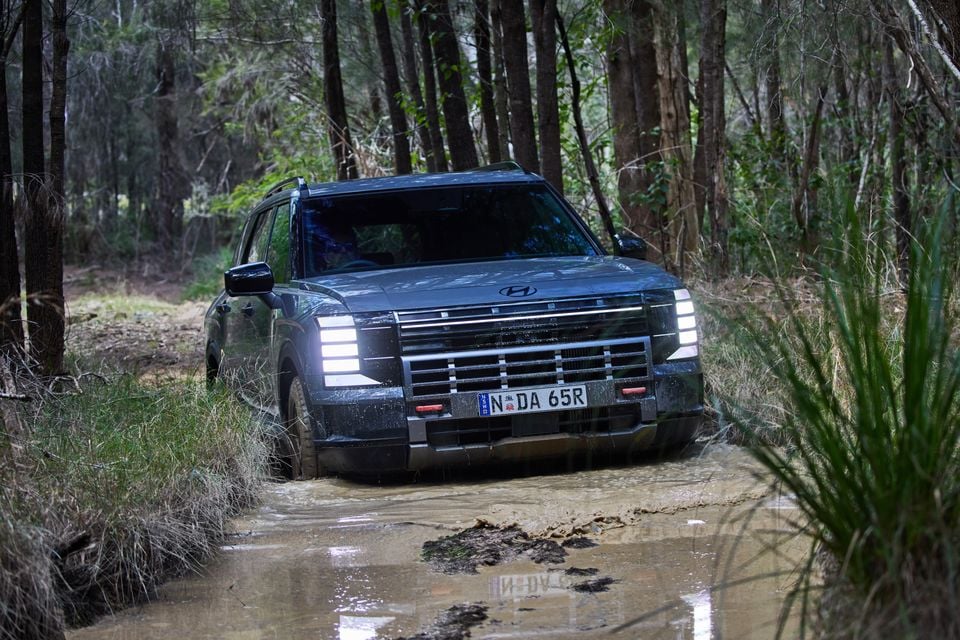 A dark grey Hyundai Palisade XRT Pro SUV driving through a muddy water crossing on a bush track, seen from the front