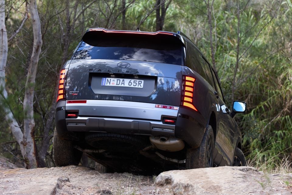 Rear view of a Hyundai Palisade XRT Pro SUV climbing over rocks on an off-road trail surrounded by Australian bush