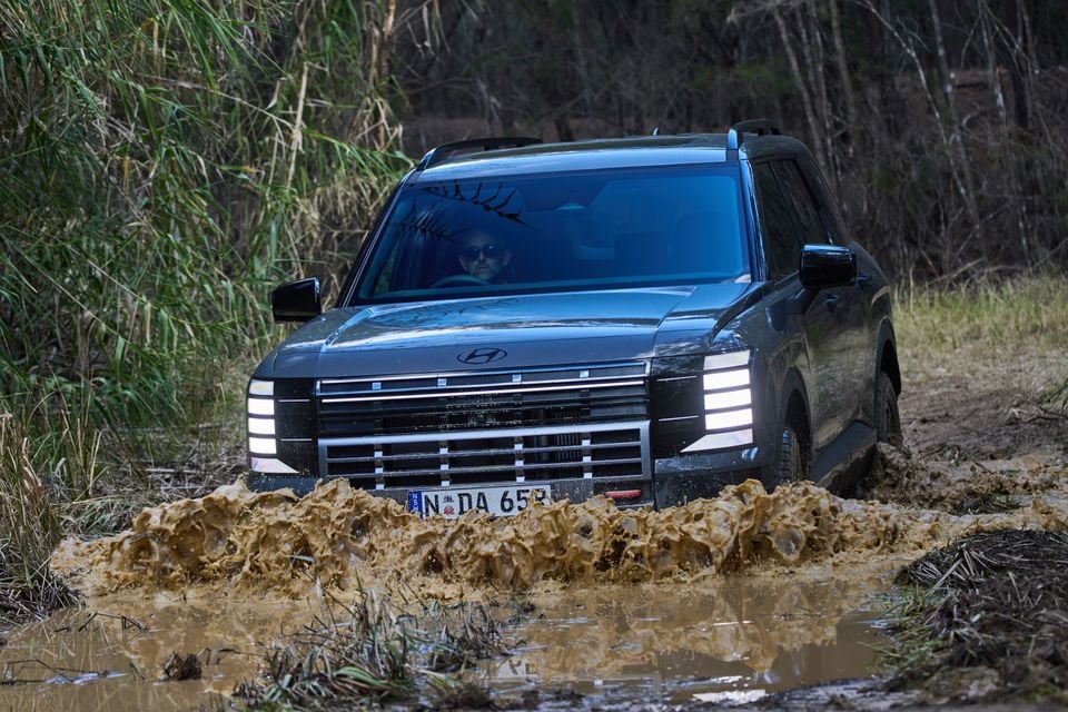 A Hyundai Palisade SUV driving through a muddy off-road track, seen from the front, with mud splashing around the bumper.