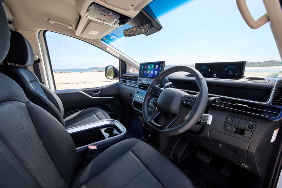 Interior of a Hyundai Staria Load Electric showing the front cabin with steering wheel, dual digital screens, centre console, and fabric seats, parked near a beach