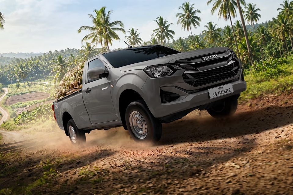 A grey Isuzu D-Max Spark single-cab ute carrying a load of coconuts, driving along a dusty dirt track through a palm plantation