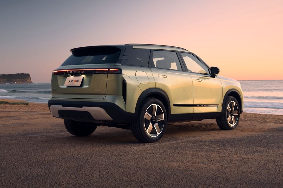 A Jaecoo J7 PHEV SUV in light green, seen from the rear three-quarter angle, parked near a beach at sunset.