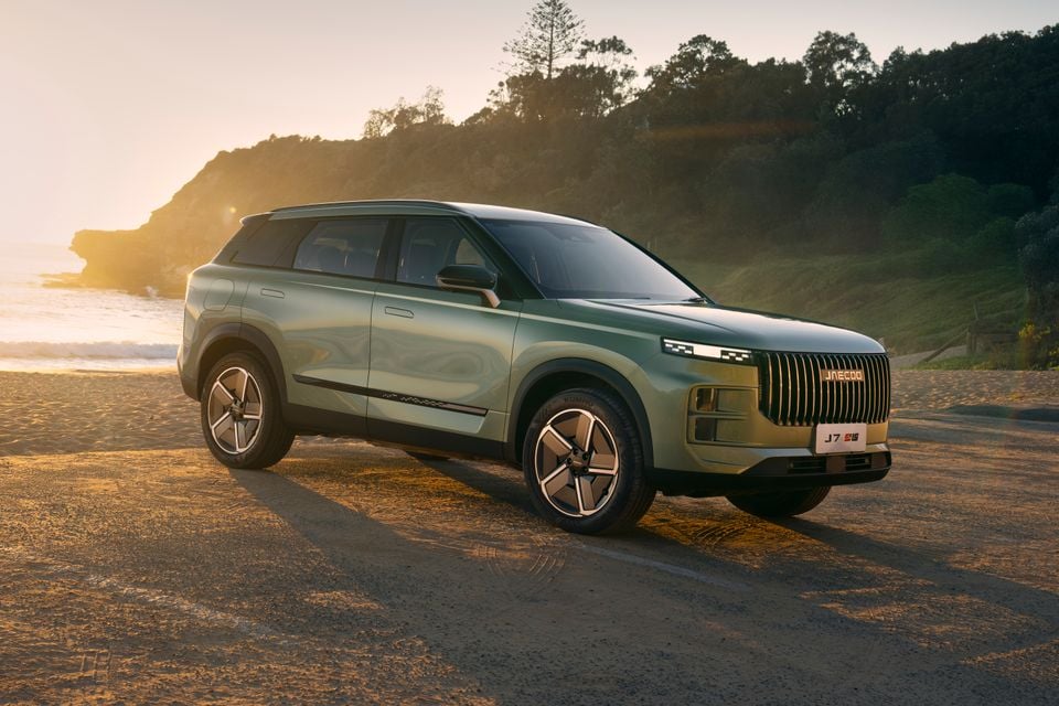 A green Jaecoo J7 SHS SUV parked on a sandy beach at sunset, seen from the front-side angle