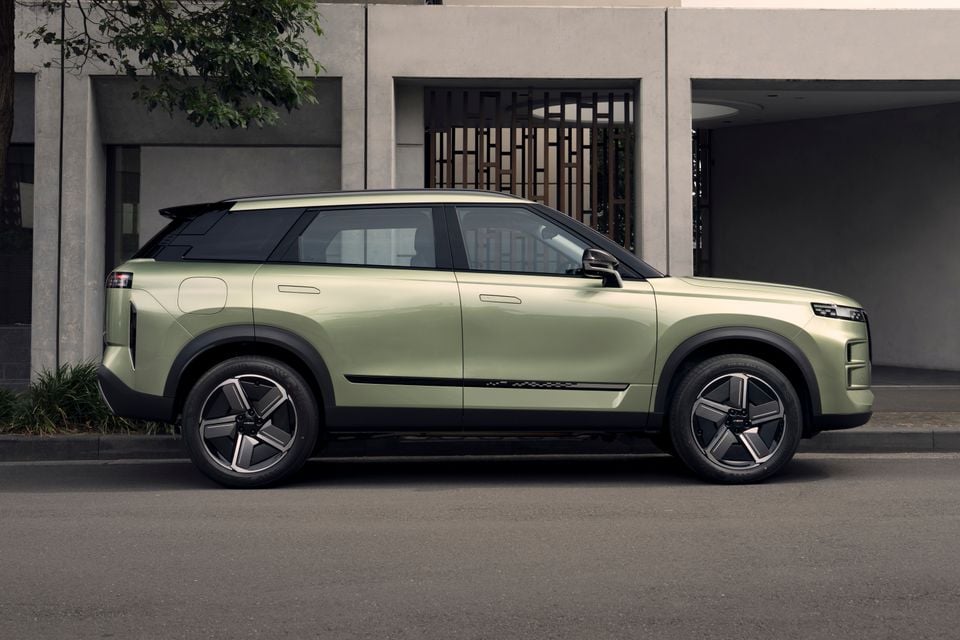 A light green 2027 Jaecoo J7 SHS SUV seen from the side, parked on a street beside a modern building