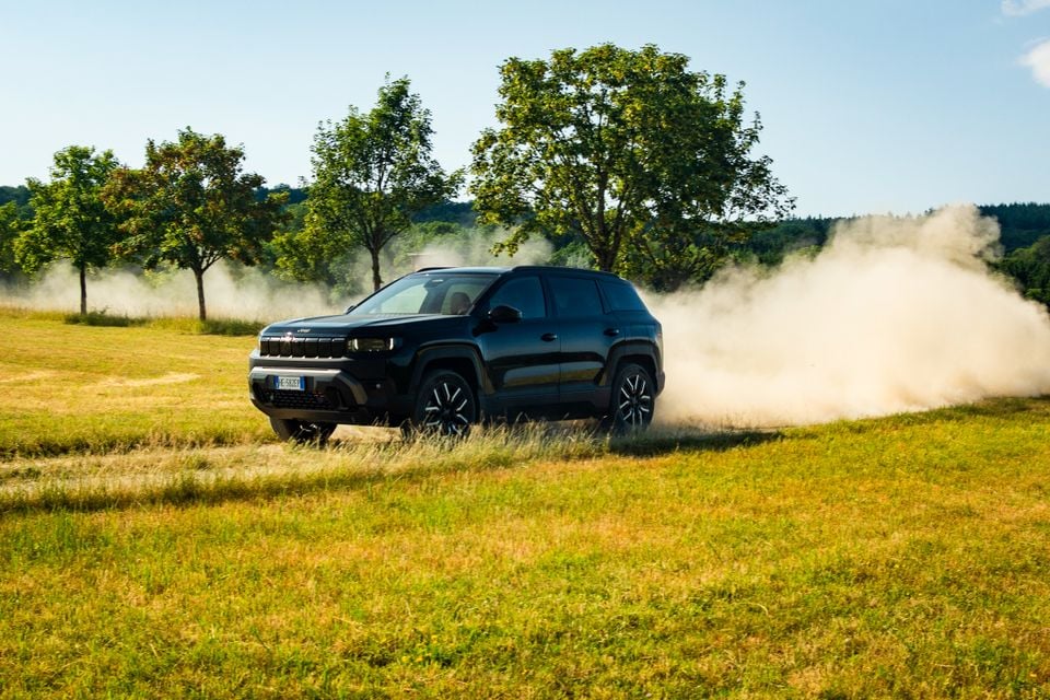 A black 2027 Jeep Compass SUV driving across a grassy field, kicking up a cloud of dust, with trees in the background.