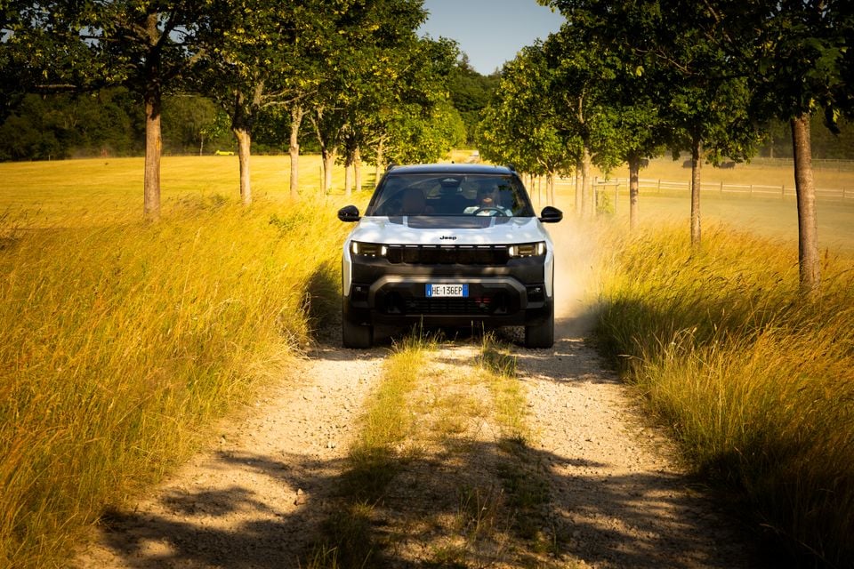 A white Jeep Compass SUV driving along a narrow dirt track through golden grassland and trees, seen from the front