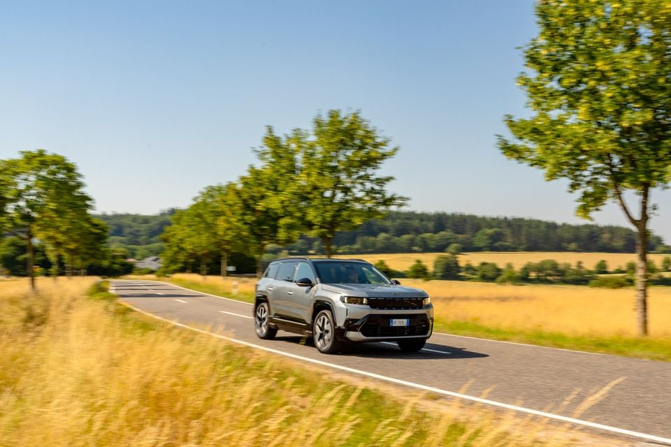 A silver Jeep Compass SUV driving along a rural tree-lined road surrounded by golden fields on a sunny day, seen from the front-side angle.