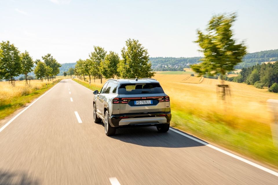 A Jeep Compass SUV seen from the rear while driving along a rural road surrounded by fields and trees