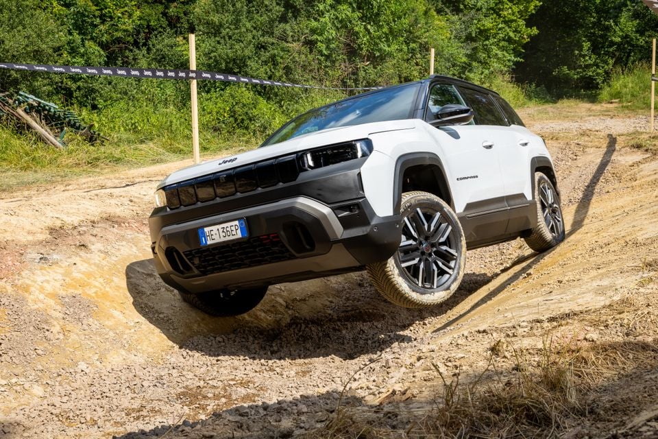 A white Jeep Compass SUV navigating a rough off-road dirt track, seen from the front-side with one wheel lifted off the ground