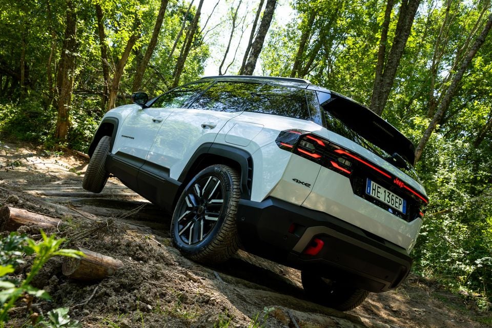 A white Jeep Compass 4xe SUV tackling a rocky off-road trail in a forest, seen from the rear three-quarter angle