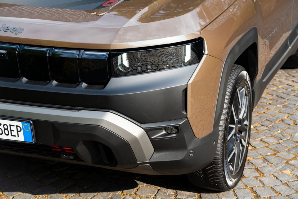 Close-up of the front corner of a brown Jeep Compass SUV, showing the headlight, grille, bumper and front wheel on a cobblestone surface