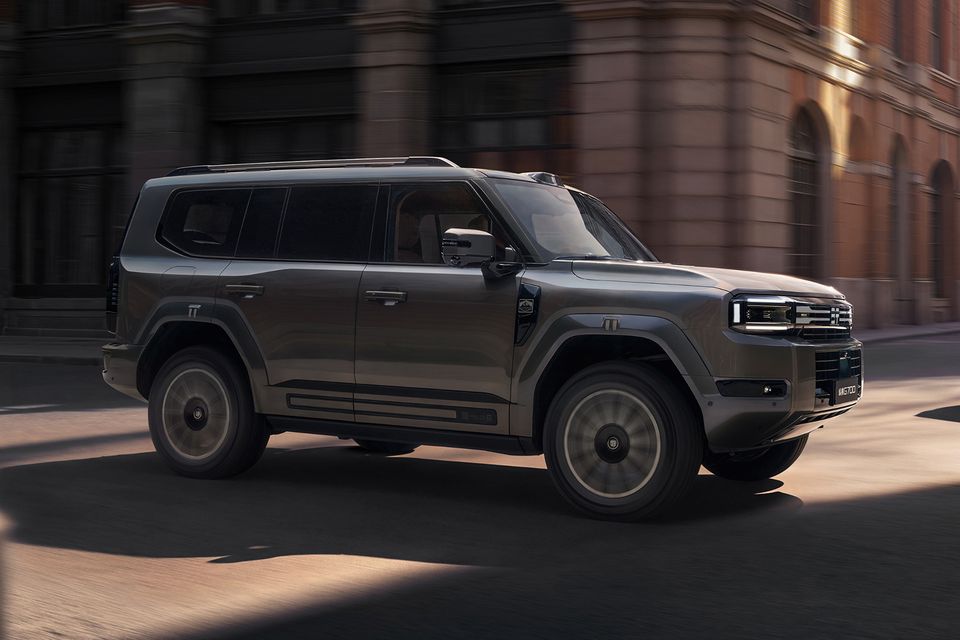 A dark grey 2027 Jetour G700 SUV driving along a city street past brick buildings, seen from the front side