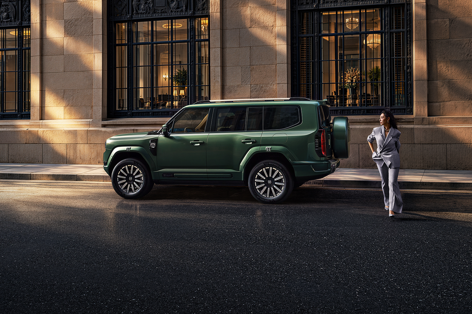 Green 2027 Jetour G700 SUV seen side-on, parked on a street outside a stone building as a woman in a grey suit walks nearby