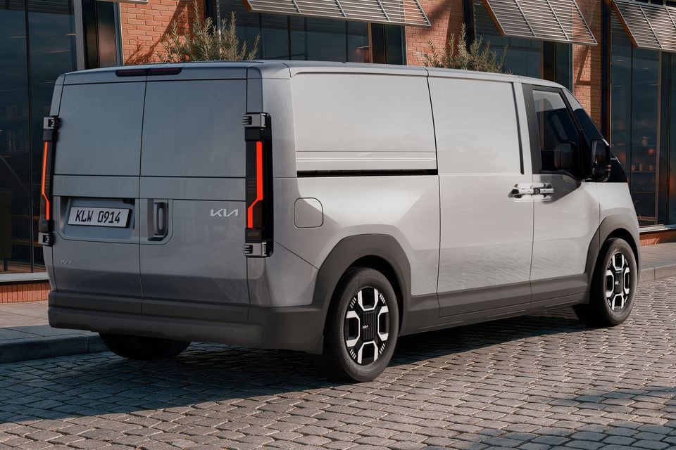 A grey Kia PV7 electric van seen from the rear three-quarter angle, parked on a cobblestone street outside a modern building