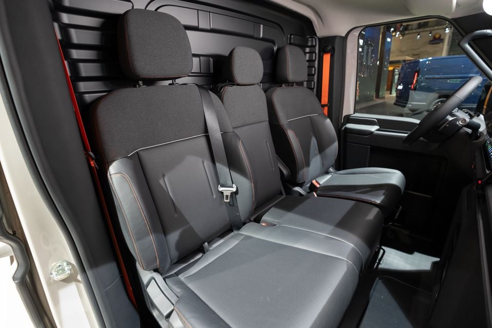 Interior view of a van's front cabin showing three-seat bench and individual seat configurations with dark fabric and orange stitching upholstery, seatbelts visible, and steering wheel to the right