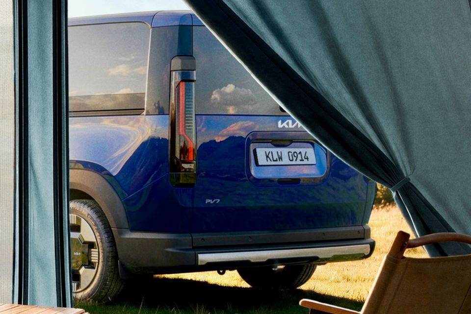 The rear of a blue Kia PV7 van parked on grass, viewed from behind a tent curtain in an outdoor camping setting.