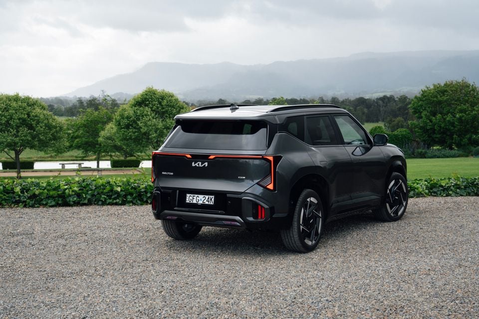 A grey 2027 Kia Seltos GT-Line hybrid SUV parked on gravel, seen from the rear with green gardens and misty mountains behind