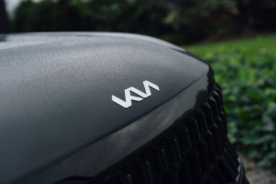 Close-up of the Kia logo on the rain-speckled dark grey bonnet of a 2027 Kia Seltos GT-Line hybrid, above its black grille