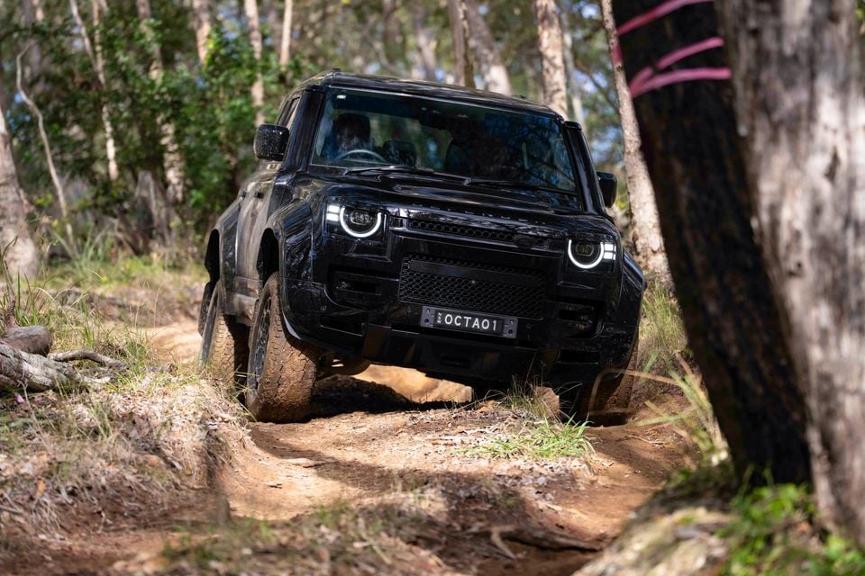 A black Land Rover Defender navigating a rugged dirt track through bushland, seen from the front