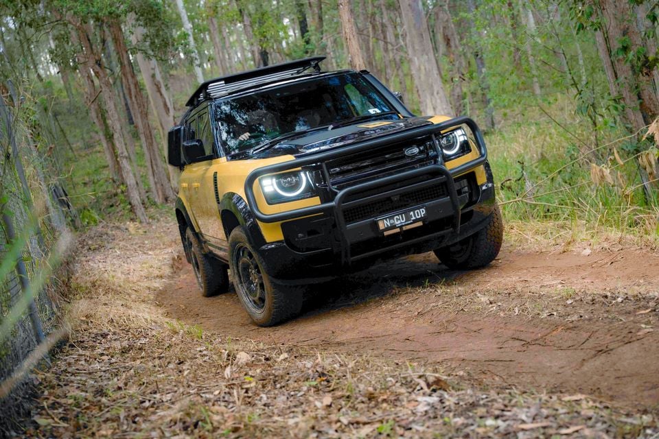 A yellow Land Rover Defender 90 with a black bull bar and roof rack driving along a dirt track through a bush setting in Australia