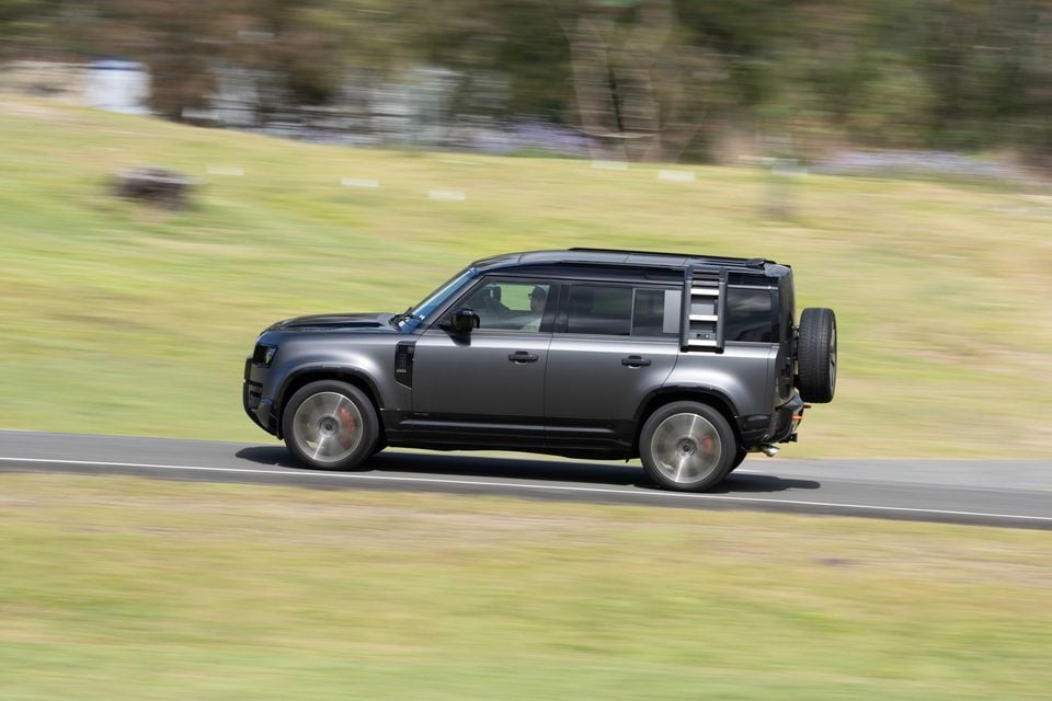 A dark grey Land Rover Defender 110 SUV driving along a road, seen from the side, with a rear-mounted spare wheel and roof ladder visible