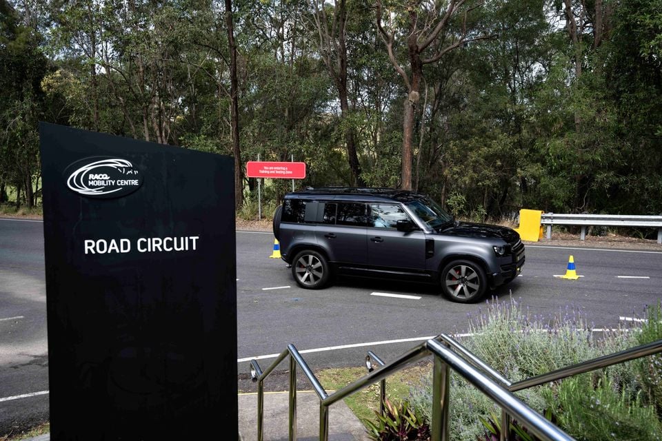 A dark grey Land Rover Defender 130 SUV driving on the road circuit at the RACQ Mobility Centre, seen from the side