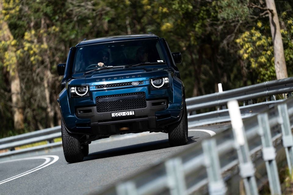 A blue Land Rover Defender SUV driving towards the camera on a winding road surrounded by trees