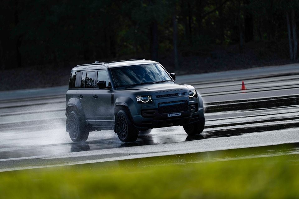 A dark grey Land Rover Defender 130 SUV driving on a wet track, seen from the front and side