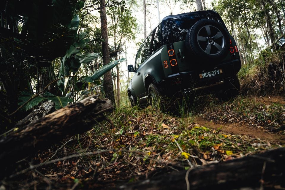 A dark green Land Rover Defender 90 seen from the rear, navigating a steep off-road trail through dense bushland in Australia