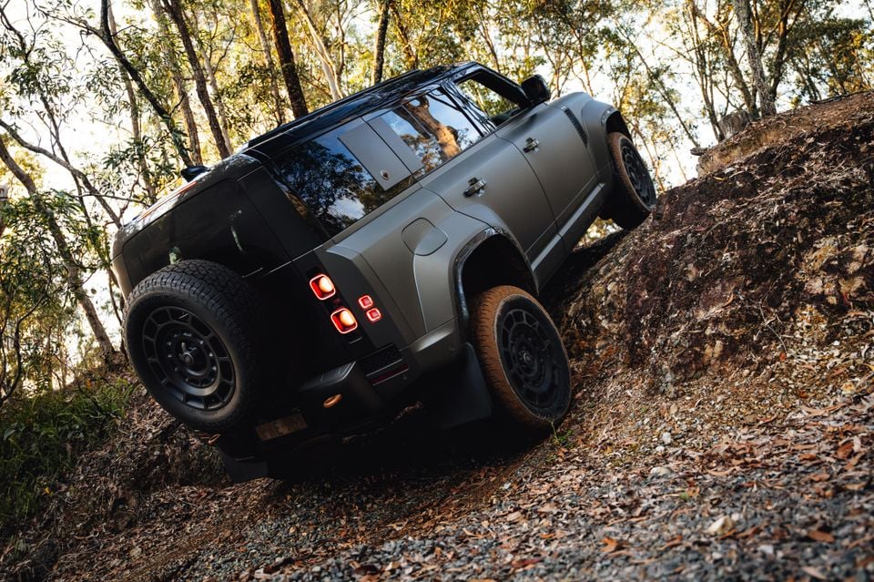A dark grey Land Rover Defender SUV climbing a steep rocky off-road trail through Australian bush, seen from the rear three-quarter angle