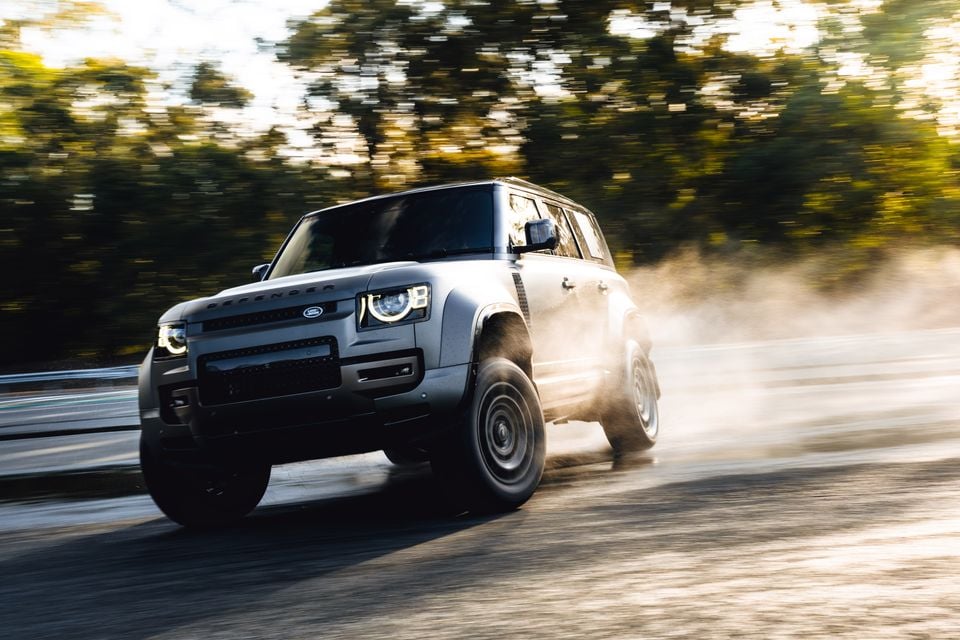 A silver Land Rover Defender SUV driving at speed on a wet road, kicking up dust and water, seen from the front-side angle with trees blurred in the background