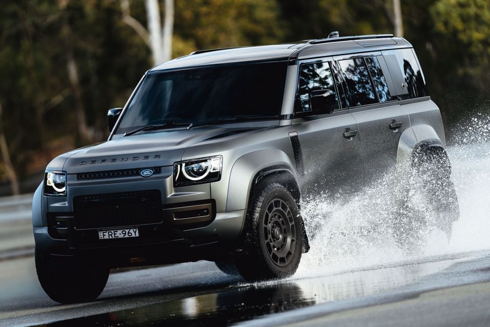 A grey Land Rover Defender 110 SUV driving through water, sending a dramatic splash, seen from the front-side angle on a wet road surrounded by trees.