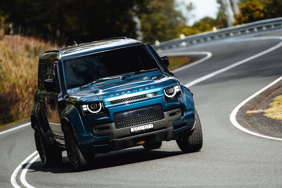 A blue Land Rover Defender SUV seen from the front while driving along a winding road
