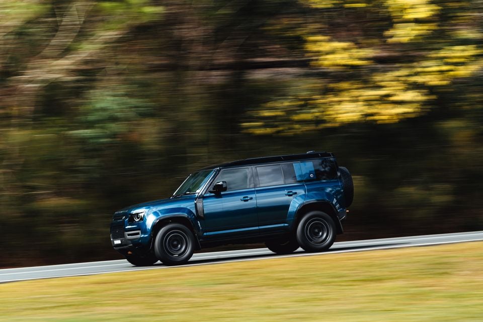 A blue Land Rover Defender 90 driving along a road, seen from the side with a blurred tree backdrop