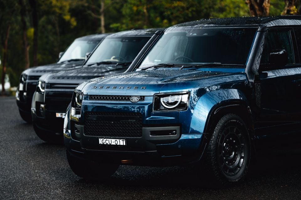 Three Land Rover Defenders lined up on a wet road, with a blue Defender in the foreground seen from the front, surrounded by trees
