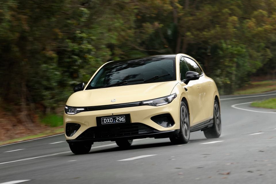 A pale yellow 2027 Leapmotor B05 hatchback with a black roof driving along a winding tree-lined road, seen from the front.
