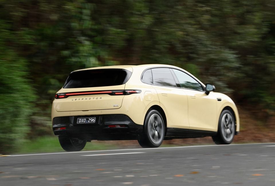 A yellow 2027 Leapmotor B05 hatchback driving on a country road, seen from the rear and side with a blurred forest background