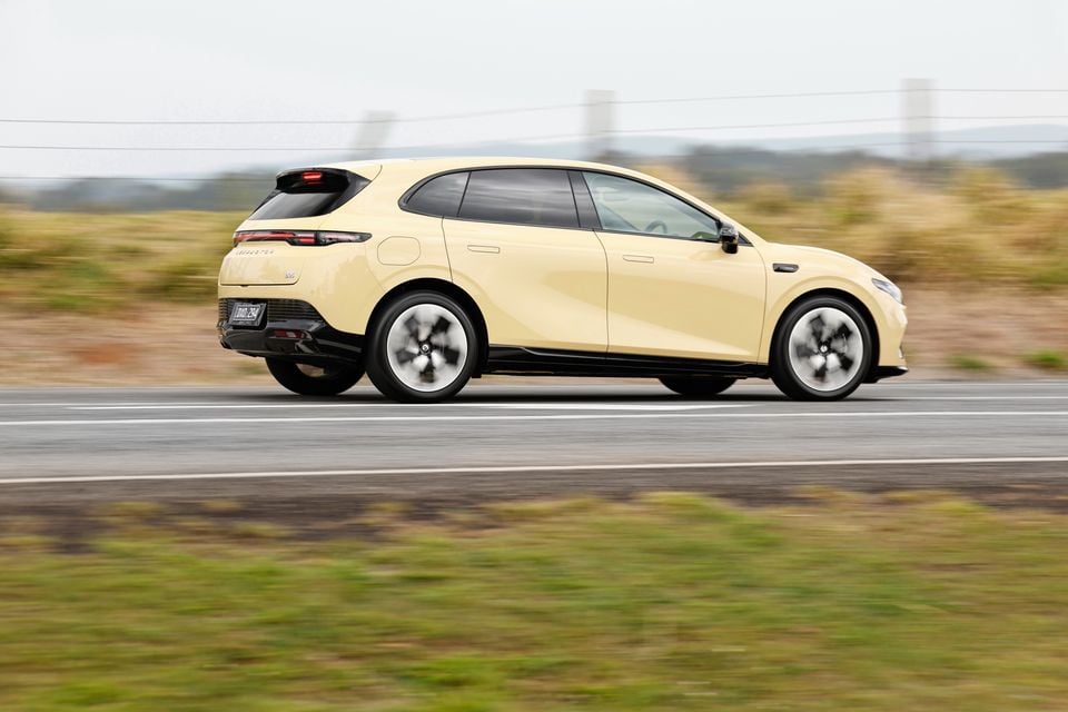 A pale yellow 2027 Leapmotor B05 hatchback driving along a country road, seen from the side and slightly behind