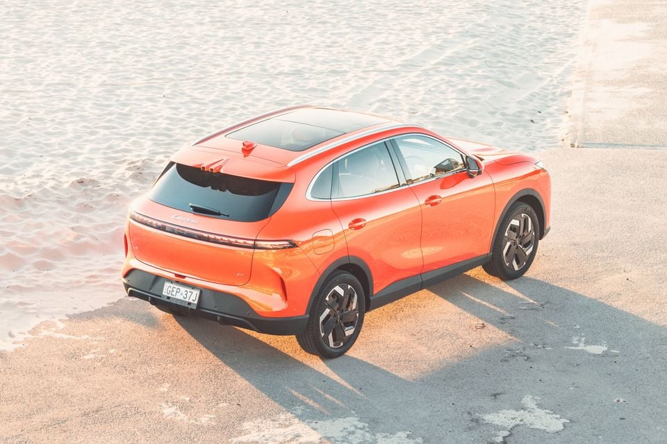 An orange 2027 Lepas L6 Signature EV SUV seen from above and behind, parked on a concrete path beside a sandy beach