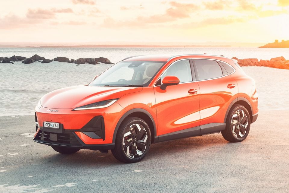 An orange 2027 Lepas L6 Signature electric SUV parked on a beachside path at sunset, seen from the front left side