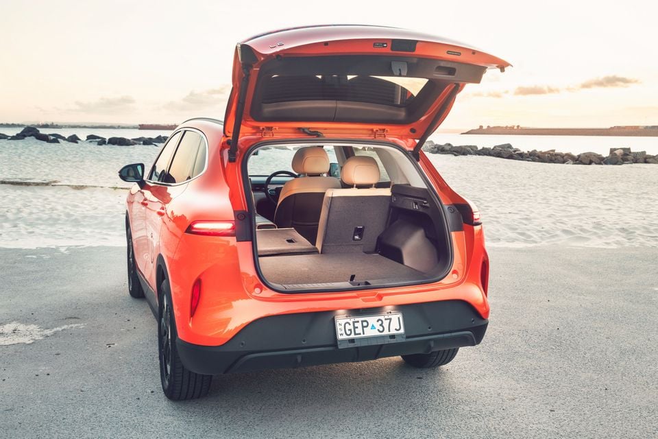 An orange 2027 Lepas L6 electric SUV seen from the rear with its tailgate open, showing the boot and a folded rear seat, parked beside a beach